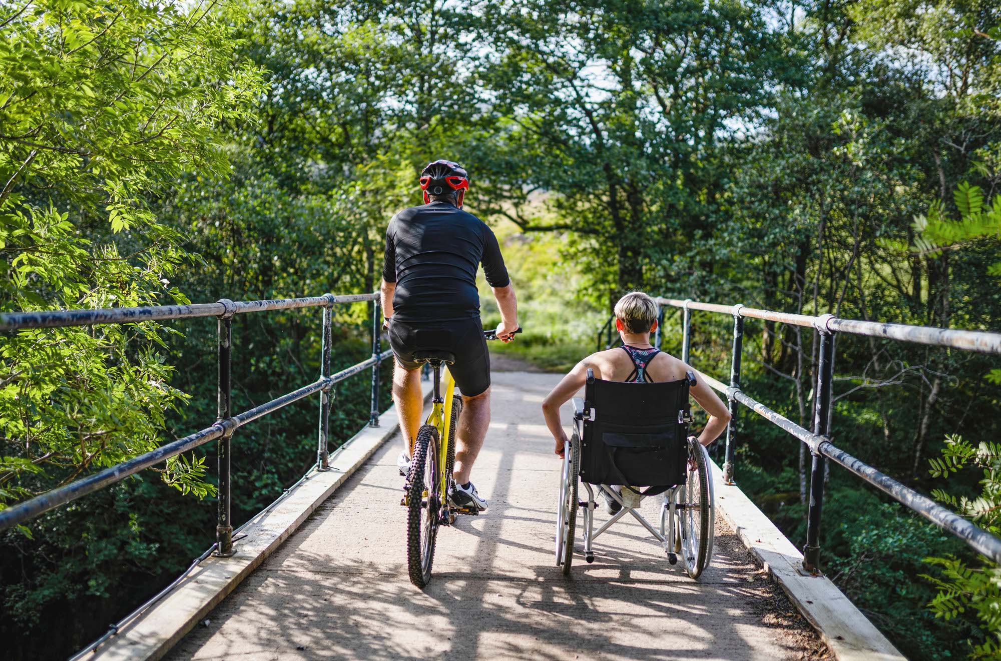 Eine Frau im Rollstuhl, begleitet von einem Freund auf dem Fahrrad, fährt über eine Holzbrücke in einem grünen Wald