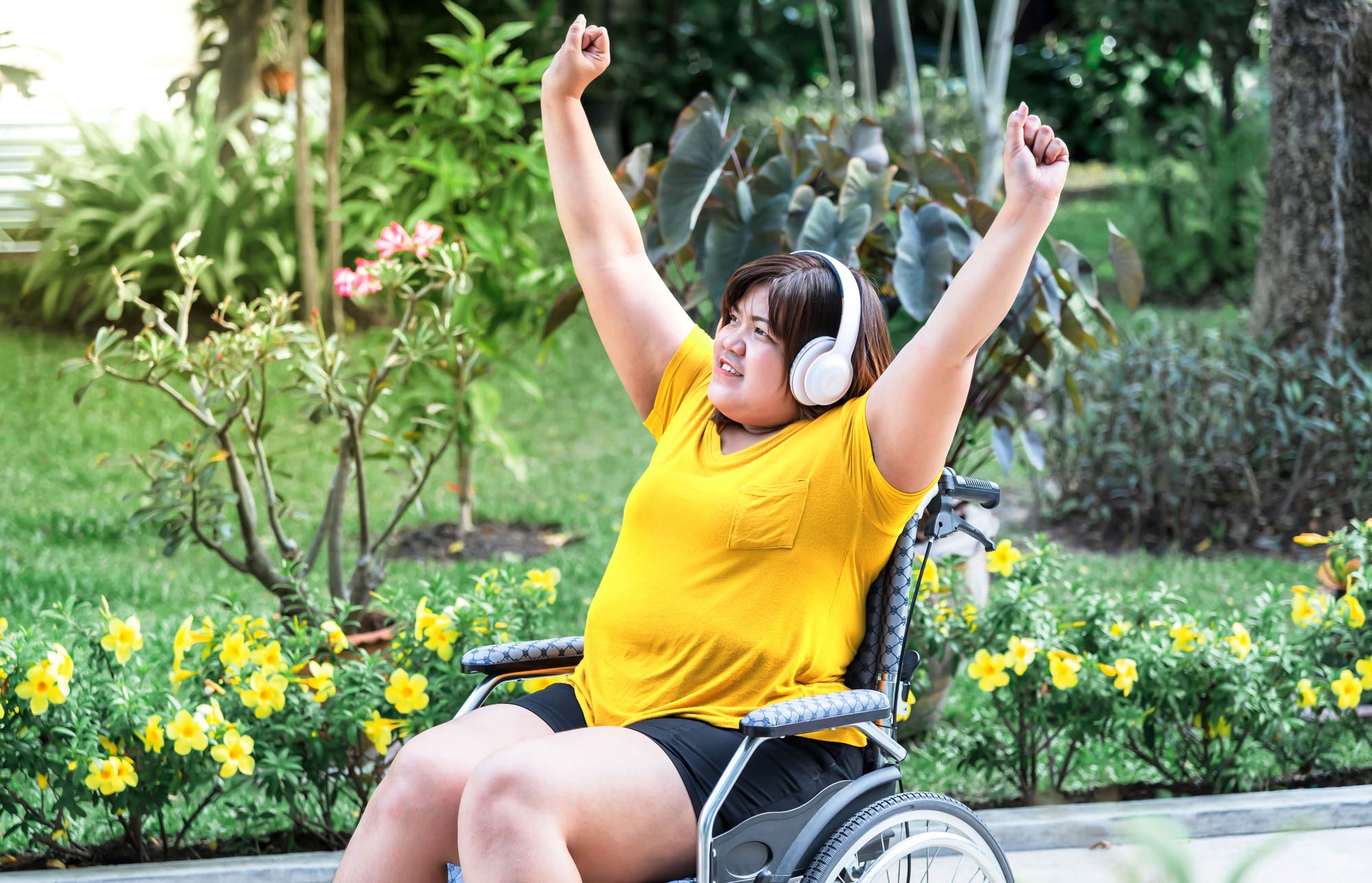 Junge Frau sitzt in einem Rollstuhl und trägt Kopfhörer, um Musik zu hören, entspannt im Garten
